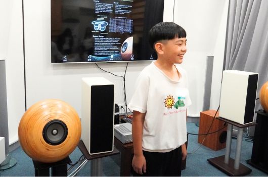 年輕男子穿著白色T恤，站在現代科技展示室內。身旁展示有木製球形揚聲器、黑色音箱和電子設備。身後牆上掛著大型顯示屏，播放技術資訊。藍色地板和灰色窗簾營造專業展覽環境。
