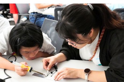 兩名女性在工作坊中專注地進行電子設備組裝或修理。其中一人穿著黑色外套，正用工具操作電子零件，另一人穿著白色上衣在旁協助。工作台上擺放著各種電子元件和工具，背景有其他參與者。