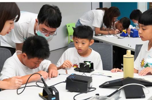 課堂上，多名穿著白色制服的學生圍坐在桌邊，專注於組裝電子機器人。他們正在操作焊槍和電路板，進行科技教育實驗。教室背景為現代化教學環境，展現STEM教育的實踐場景。