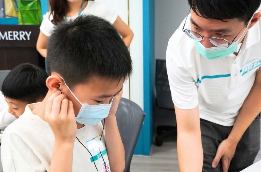 醫護人員為學童進行健康檢查，小男孩戴著口罩坐著接受檢查，醫生穿著白袍和口罩在旁邊協助，背景有其他學童等候，室內環境明亮整潔。