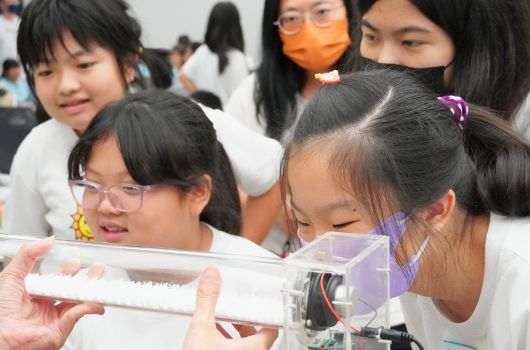一群穿著白色制服的學生在科學實驗室中認真觀察手中的電子裝置。學生們戴著彩色眼鏡和髮飾，聚集在一起查看展示板上的科學儀器和實驗材料，參與互動式的教育活動或科學展示。