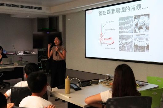 演講者於會議室進行簡報，投影螢幕顯示有關環境議題的內容，包含圖表及照片。聽眾坐在桌前專注聆聽，場景為室內學術或企業演講環境。