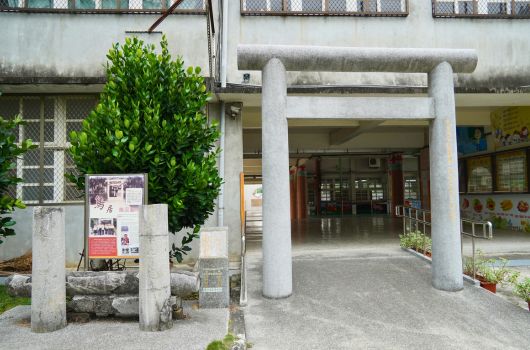 一棟老舊建築的入口處，有兩根白色圓柱支撐著門廊。左側柱子旁放著一塊告示牌，上方有綠色盆栽植物。入口深處可見玻璃門和室內空間，牆面貼有海報和裝飾。整體呈現樸素的公共設施或社區中心的外觀。