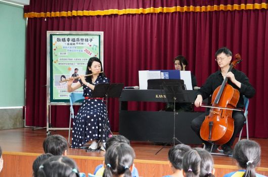 舞台上三位音樂家正進行室內樂表演，左方演奏小提琴，中間在鋼琴前，右方演奏大提琴。舞台後方豎立宣傳海報，觀眾席坐滿後排觀眾正欣賞演出。