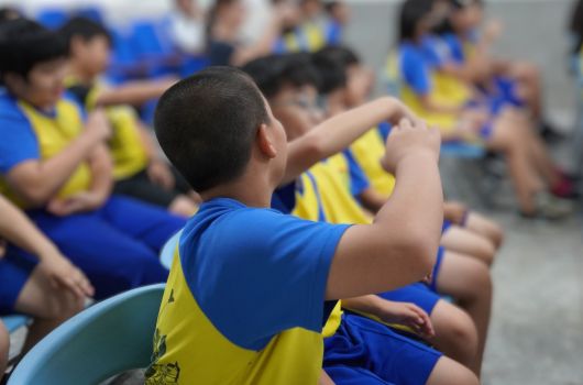 一群穿著藍黃相間運動服的兒童坐在地上，舉起雙臂參與集體活動。焦點在前景一名兒童身上，他穿著藍黃色衣服，雙臂向上伸展。背景有多名兒童也做出相同動作，顯示這是一場組織有序的群體運動或課程。
