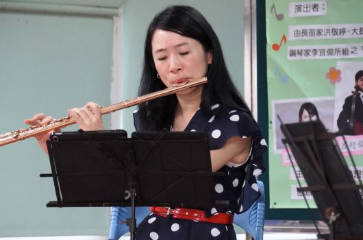 一位穿著黑色波點上衣的女性在教室內吹奏長笛，面前放置樂譜。背景為綠色牆面和教學海報，右側可見其他學生。這是一場音樂表演或教學活動的現場。