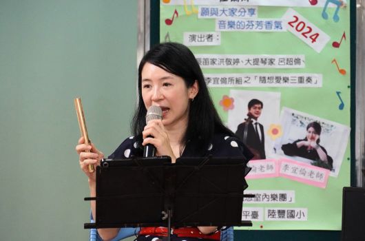 一位穿著黑色服裝的女性站在麥克風前，手持筷子，身後是一個綠色海報，上面寫著「2024嶺東大家分享音樂的芬芳香氣」，展示各項表演者資訊與活動內容。