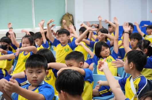 一群穿著黃藍色運動服的小學生在室內聚集，舉起雙手做出歡樂的姿勢。孩子們聚在一起參與團體活動或課程，展現活力與參與度。背景是白色牆壁和綠色黑板。