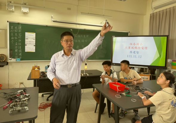 教室內，一位戴眼鏡的男性教師舉起手機向學生展示內容，身後黑板和螢幕顯示教學資訊。四位學生坐在課桌前，桌上放置機器人和電子零件，認真聽講。這是一堂科技或機器人教育課程。