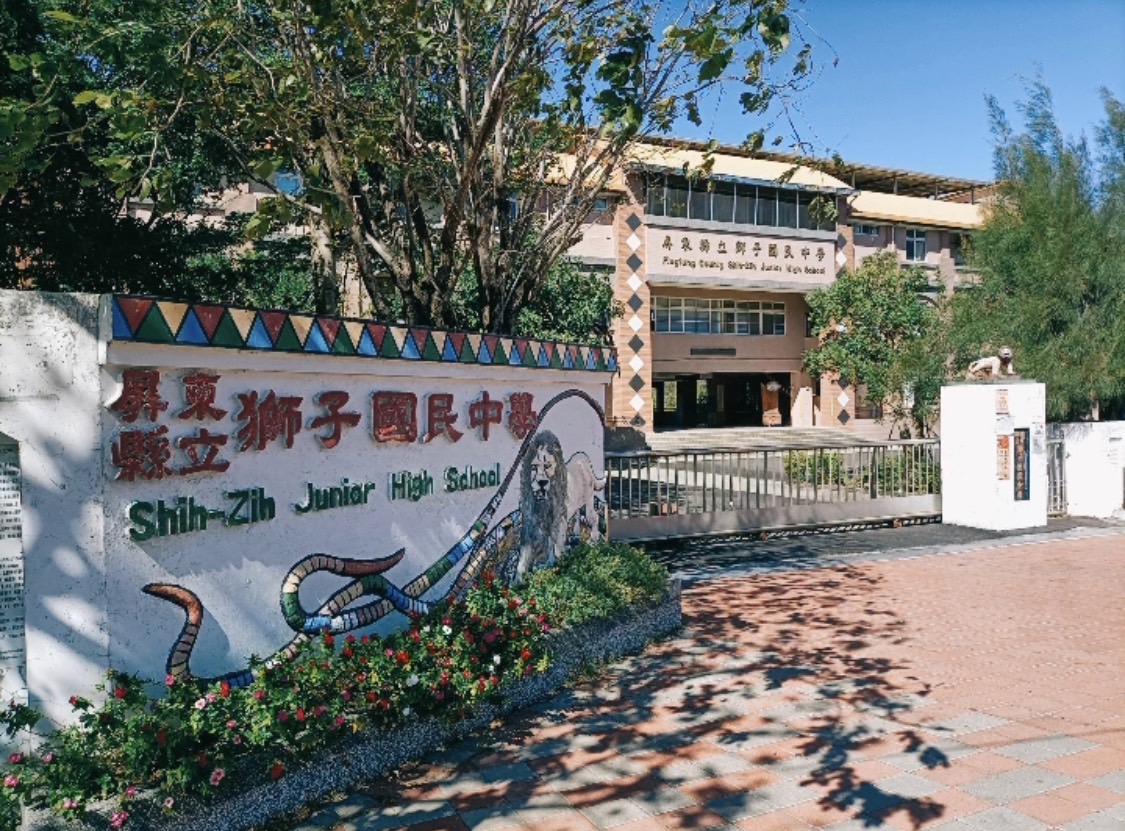 獅子國中校園入口，白色圍牆上繪有校名「獅子鄉立獅子國民中學」與彩色蛇形圖案。背後是紅磚校舍，門前鋪設紅色地磚，周圍種植樹木花卉，晴朗天氣下呈現校園風貌。