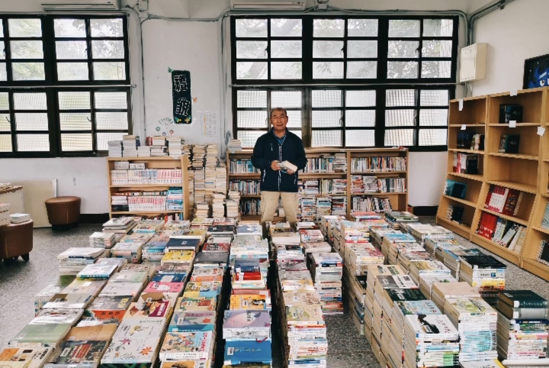 一名男性在圖書館內站立，身前地面排滿數百本整齊堆疊的書籍，展示了豐富的藏書。背景為木質書架、大型黑框窗戶和教室設施，呈現圖書館或校園閱讀推廣的場景。