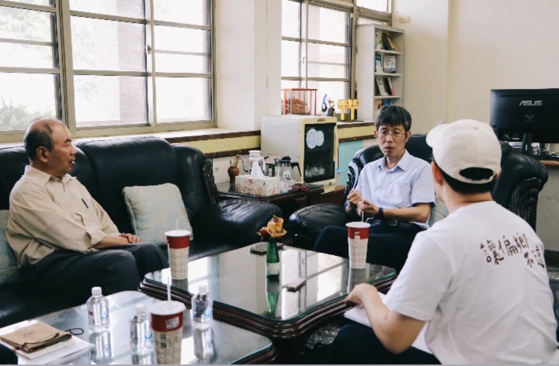 三位男性在辦公室內進行會議討論，坐在黑色沙發旁。桌上放有飲料和水瓶。背景為大窗戶、書櫃、電腦螢幕和各式裝飾品，呈現專業的工作環境。