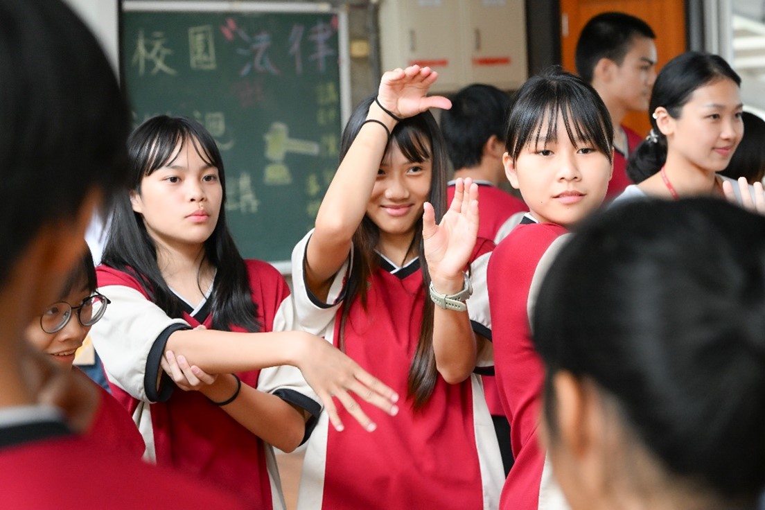 一群穿著紅白色校服的學生在教室內進行團隊活動，他們圍成一圈，將手臂放在中央堆疊在一起，展現團隊合作精神。背景可見綠色黑板和教室環境。