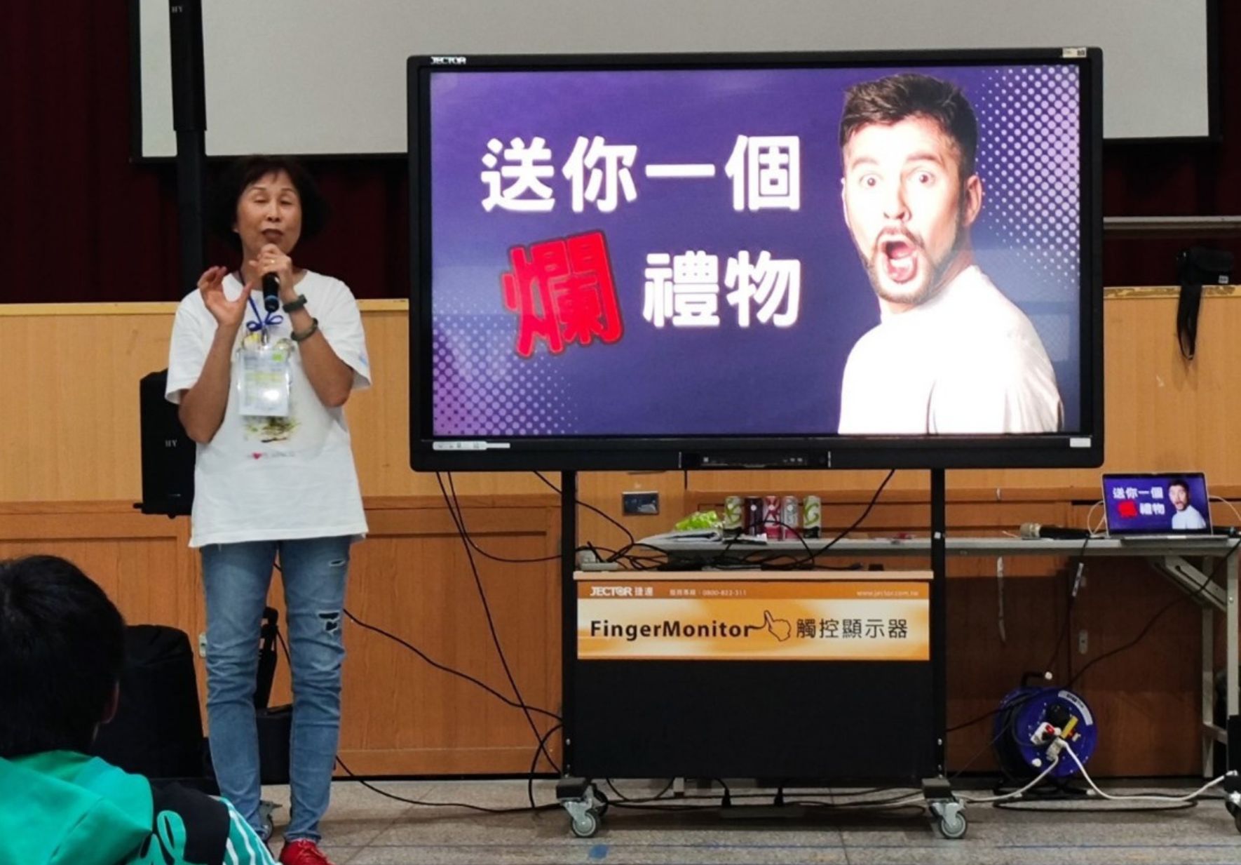 一位女性講者在教室內拿著麥克風進行簡報。身後大螢幕顯示中文文字「送你一個驚禮物」，搭配一名男性的驚訝表情影像。講台下方可見FingerMonitor觸控顯示器和其他簡報設備。