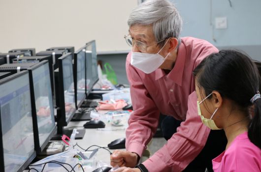 一名戴著口罩的年長男性講師在電腦教室中，彎身指導一位穿著粉紅色衣服、戴著眼鏡的女性學員操作電腦。背景可見多台桌上型電腦排列在教室內。