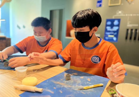 兩名穿著橙色制服、戴著口罩的學生在教室內進行陶藝課程。右邊學生在藍色墊子上操作陶土，左邊學生也在進行類似活動。桌上放置陶藝工具和材料，背景有教室設備和海報。