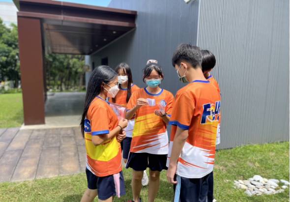 四名穿著橙色運動衣的學童在戶外草地上聚集，圍成一圈進行團隊活動。背景是現代建築物和綠色草坪，展現校園運動或團隊建築活動的情景。