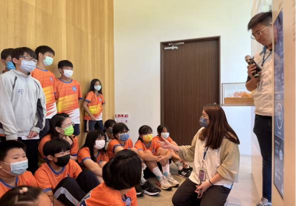 室內教室場景，一位穿著米色外套的女性講師正面向坐在地上穿著橙色制服的兒童進行互動教學，另有男性講師站在一旁操作投影設備，現場約有十多名兒童戴著口罩參與活動。