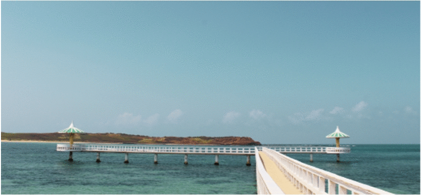 一條現代化的白色跨海大橋橫跨碧綠色海面，橋身兩端各有金色尖頂塔樓。遠方可見褐色陸地與清澈蔚藍天空，天氣晴朗，幾朵白雲點綴其中。