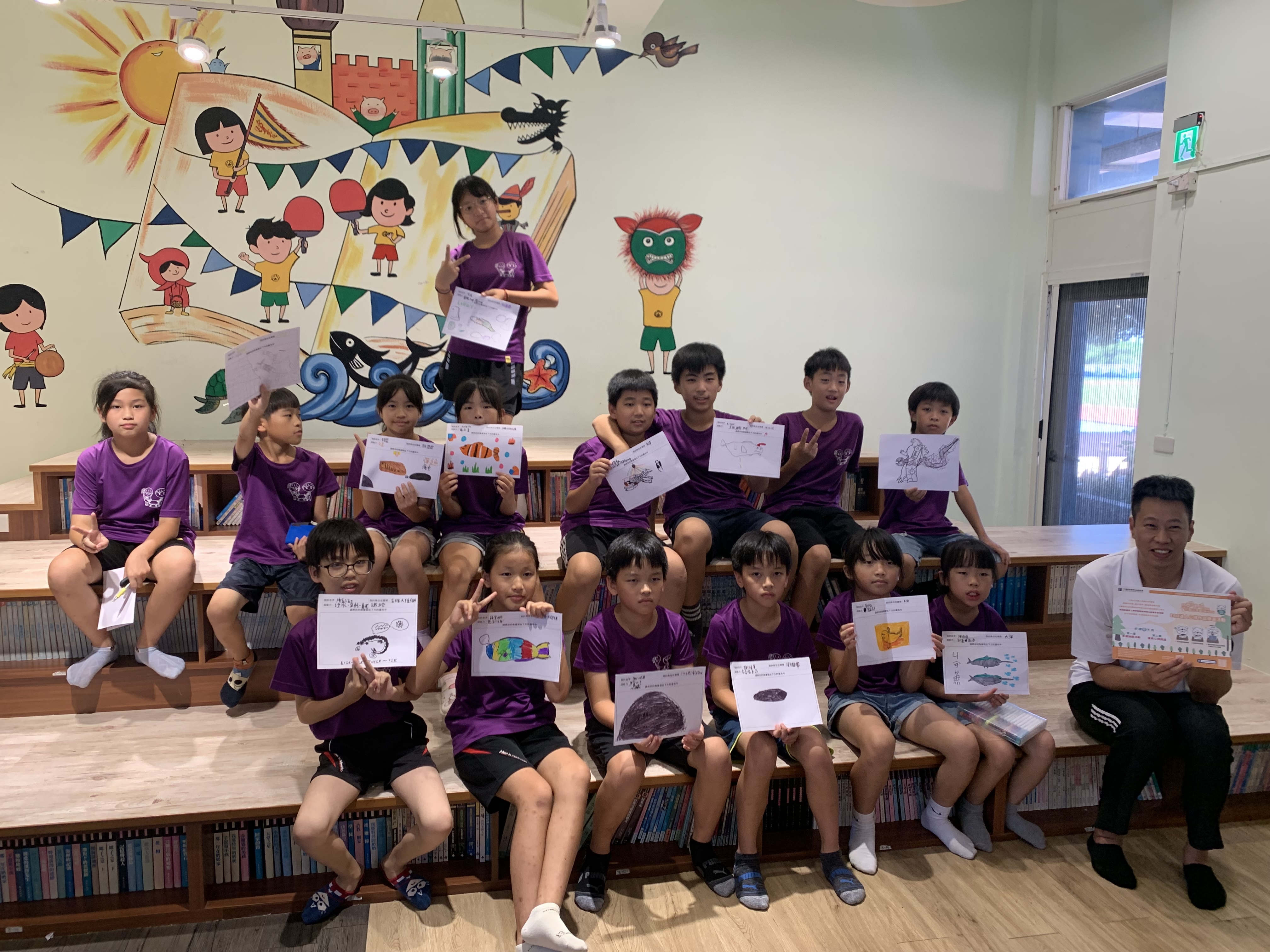 一群穿著紫色制服的學童在圖書館內展示他們的繪畫作品。背景牆上有色彩繽紛的兒童主題壁畫，包含帆船、城堡和卡通人物。學童們坐在書架前，開心地舉起各自創作的圖畫，展現美術課的成果。