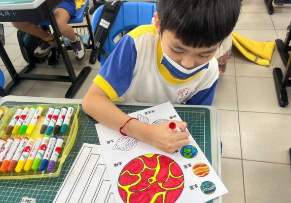 小學生穿著藍黃色校服，坐在教室裡用彩色筆進行美術創作。學生正在為行星主題的著色圖上色，桌上擺放著整齊排列的彩色筆和綠色墊板。周圍有其他同學也在進行相同的美術課程活動。