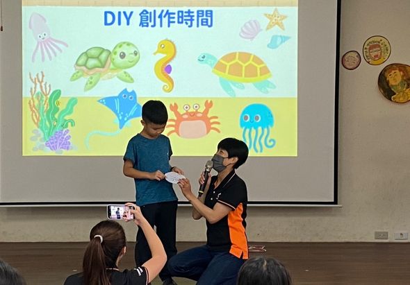 教室內進行DIY創作時間活動，講師在投影幕前向學生示範海洋主題手工藝，幕上展示海龜、海馬、螃蟹、水母等海洋生物插圖。學生坐在地上觀看，有人用手機錄影記錄過程。