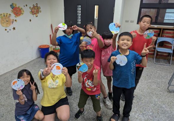 一群小學生在教室內展示他們製作的彩色紙盤藝術作品。約十名穿著各色衣服的孩童開心地舉起手中的裝飾紙盤，牆上貼著相似的黃色和橘色紙盤裝飾，背景可見教室書櫃和黑板門。