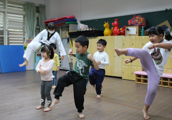 幼兒園教室內，一位教師指導約五名幼童進行跆拳道課程。幼童們穿著淺色衣服，在木質地板上擺出踢腿動作。背景有木製儲物櫃、玩具和教室布置。這是一堂身體活動課程的教學場景。