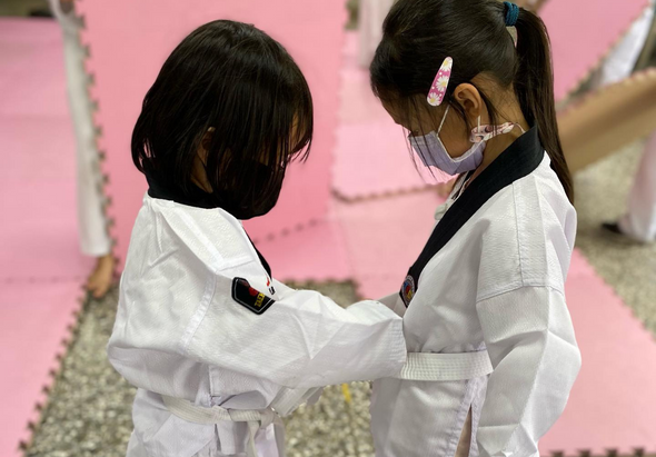 兩名穿著白色道服的小女孩在粉紅色的武術訓練墊上互相對練，其中一人戴著口罩。背景是柔和的粉紅色牆面，展現兒童武術訓練的場景。