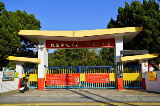 花園市花卉公園大門，白色鋼架構造，頂部黃色遮陽棚，紅色大門，門上有中文標誌和資訊牌，周圍被綠樹環繞，晴朗藍天，門前停放機車。