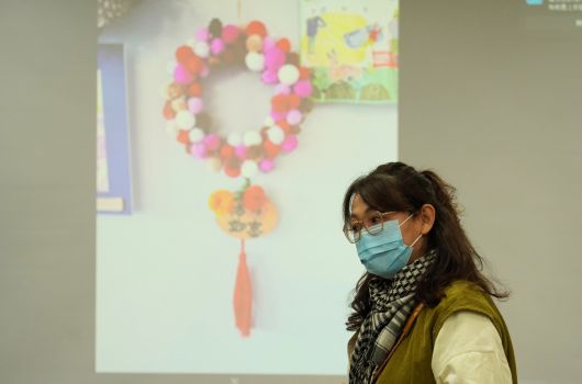 一位戴著口罩和眼鏡的女性站在牆邊，身穿黃綠色外套和黑白格紋圍巾。牆上展示著彩色的球形花環裝飾，下方掛著橙色流蘇裝飾，右側可見黃色的花卉海報。