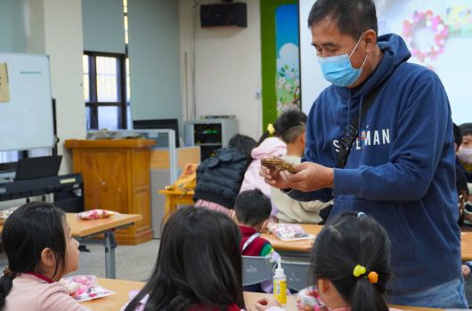 教室內，一位戴著口罩、穿著藍色外套的男性成人向坐在桌前的四名小學生展示並分發食物。學生們專注地看著他的示範，桌上擺放著食物和飲料。背景可見教室的各項設施和布置。