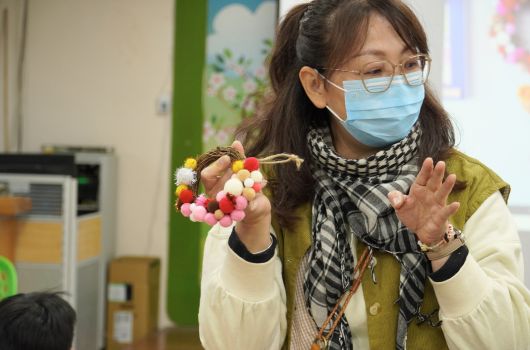 一位戴著口罩、眼鏡的女性教師，穿著黃綠色背心和格紋圍巾，在教室內舉起一個彩色毛球手工藝品。她正向學生展示美勞作品，背景是綠色牆面和教室布置。