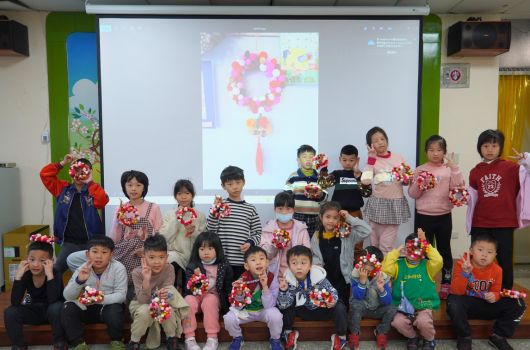 幼兒園教室內，約二十多位小朋友穿著紅白相間的服裝，手持紅色花球裝飾品排成兩排合影。背景投影幕顯示紅色花球設計圖案。場景呈現新年或春節慶祝活動的溫馨氛圍。