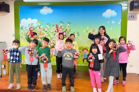 一群小學生站在教室前，背景是色彩繽紛的花卉壁畫。孩子們穿著整齊，手持粉紅色花束，展現開心的笑容。這是一場校園春季活動或才藝表演的合照。