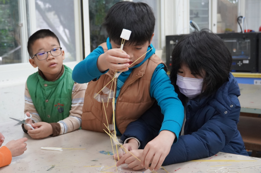 三名兒童在室內進行科學實驗，中央的男孩用手指著用繩子連接的裝置，左邊戴眼鏡的男孩和右邊戴口罩的女孩專注地觀看。桌面上放有實驗用具，背景是教室環境。