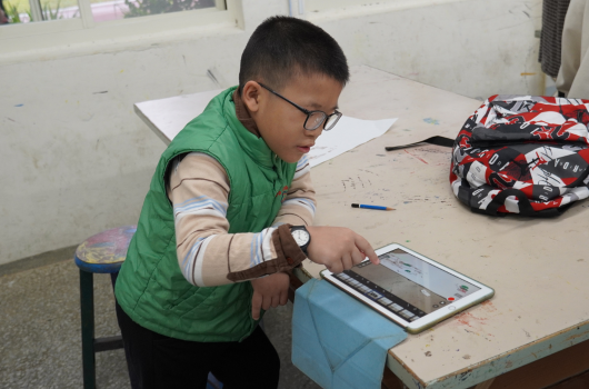 一名戴著眼鏡的男童穿著綠色外套，坐在教室裡用平板電腦進行學習。他的手臂上纏著繃帶，桌上放著紙張、筆和紅色背包，身後有白板。