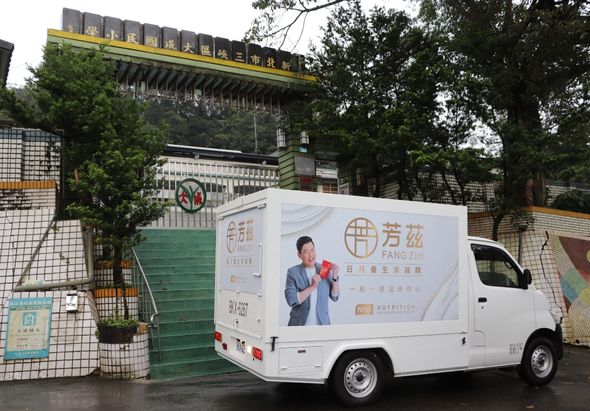 白色廂型車停靠在街道旁，車身印有「方滋」品牌標誌和一位男性形象，宣傳食品相關產品。背景為綠色鐵門和上方的黃色招牌建築，周圍環境為城市街景。