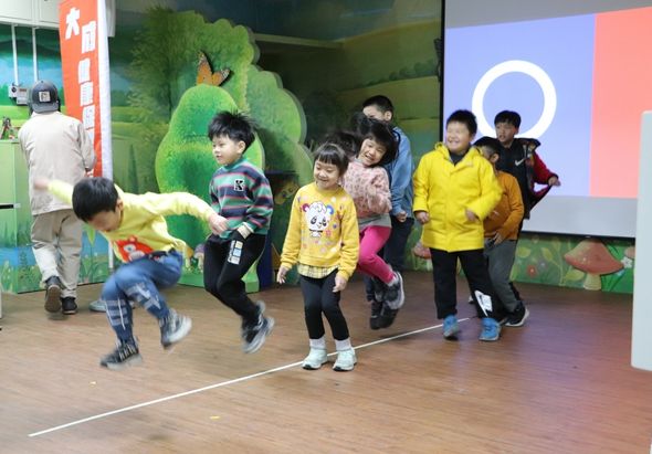 室內活動場景，多名幼童在木製舞台上跑動遊玩，背景有綠色植物布景和大型螢幕顯示。左側有成人引導，右側有家長陪同。場景色彩鮮豔，兒童穿著黃色、彩色衣服，氣氛熱鬧活潑。