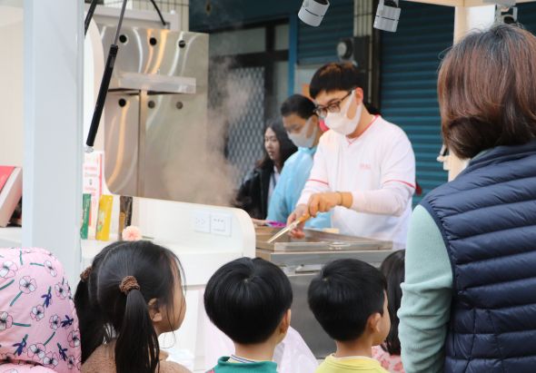 戶外美食攤位，穿著粉紅色衣服、戴口罩的工作人員正在為顧客製作食物。數名兒童和成人排隊等候，背景可見藍色鐵門和室內用餐區域。