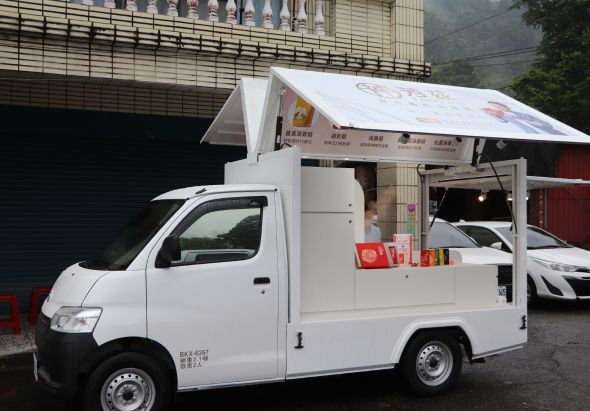 白色行動餐車停靠在街邊，配備遮陽篷和側開式服務窗口，提供街邊小食販售服務。背景為建築物和停泊的車輛，展示城市街邊商業活動的場景。