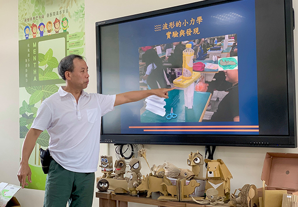 一位男性講師穿著白色短袖polo衫，指向身後的大螢幕進行教學簡報。螢幕上顯示藍色背景的投影片，標題為繁體中文，內容展示教室實景照片。講台上擺放著各式教具和模型。左側牆上貼著綠色教育海報。