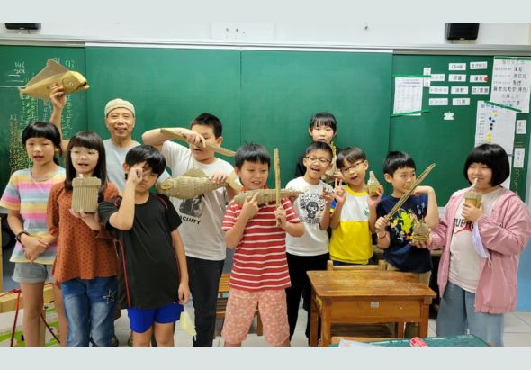 教室內一群小學生和教師在綠色黑板前合影，學生們手持木製魚形或傳統工藝品，展示學習成果。背景黑板上有教學內容和佈告欄。這是一場手工藝或自然科學的教學活動紀錄照。