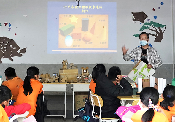 教室內，戴口罩的講師向穿著橙色制服的學童展示廢棄物回收利用教學。投影幕上顯示資源回收站製作教程，講台前擺放各種回收物品樣本，學童認真聽講。牆面裝飾環保主題布置。
