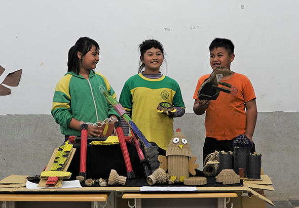 三位小學生在教室展示自製的木製機械裝置。左邊女孩穿綠黃色運動衣，中間女孩穿綠黃色制服，右邊男孩穿橙色上衣。桌上放有彩色木製零件、齒輪、滑輪等機械模型，展示科學教育活動成果。