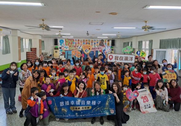 一個校園集合活動，約五十多位穿著彩色衣服的學生和老師聚集在室內場地，舉著藍色布幔和海報，共同慶祝或推廣音樂相關的校園活動。