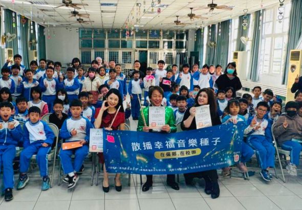學校室內集合活動，約五十位穿著藍色制服的學生與教職員合影，中央成人持藍色橫幅，上面寫著「散播幸福音樂種子」，學生們展示獲得的獎狀或證書，體現音樂教育推廣的成果。