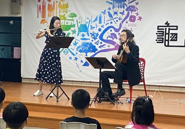 音樂教室內，一位女性小提琴手站立演奏，另一位女性坐著彈奏鋼琴伴奏。舞台背景為彩色城市插畫。觀眾坐在前排觀看表演。