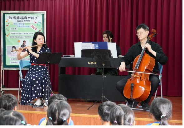 音樂表演現場，舞台上有三位音樂家，左側女性演奏長笛，中央為鋼琴，右側男性演奏大提琴。舞台背景為深紅色布幕，左側有活動海報。觀眾坐在舞台前方觀看演出。