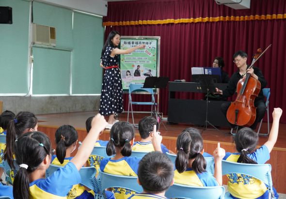 學校音樂教室內，穿著藍黃制服的小學生坐在地上聆聽音樂表演。舞臺上女講師站在海報旁，鋼琴家和大提琴手正在演奏。學生舉手踴躍參與互動，享受現場音樂教育活動。
